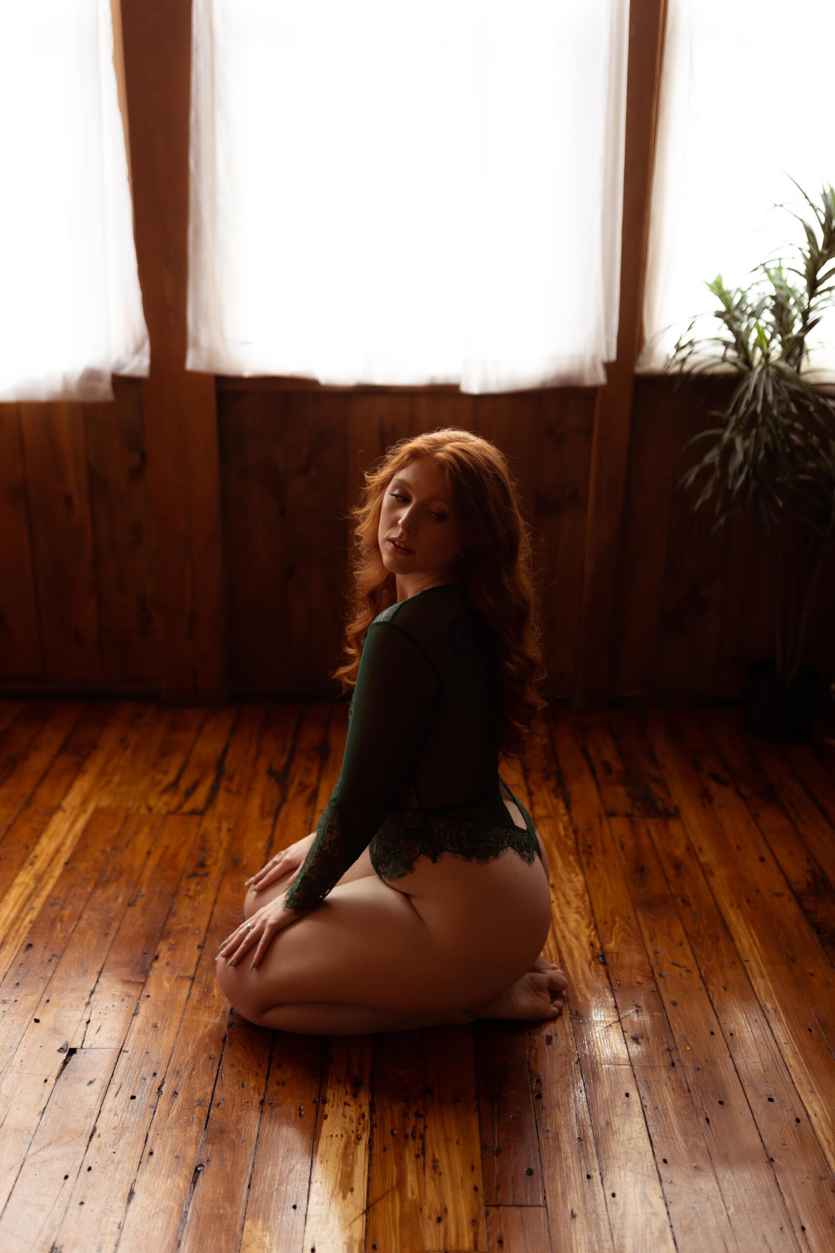 Boudoir photo of a woman with long red hair kneeling on a hardwood floor wearing a green bodysuit with windows behind her in the Boudoir by J&S Photography studio near Boston, Massachusetts