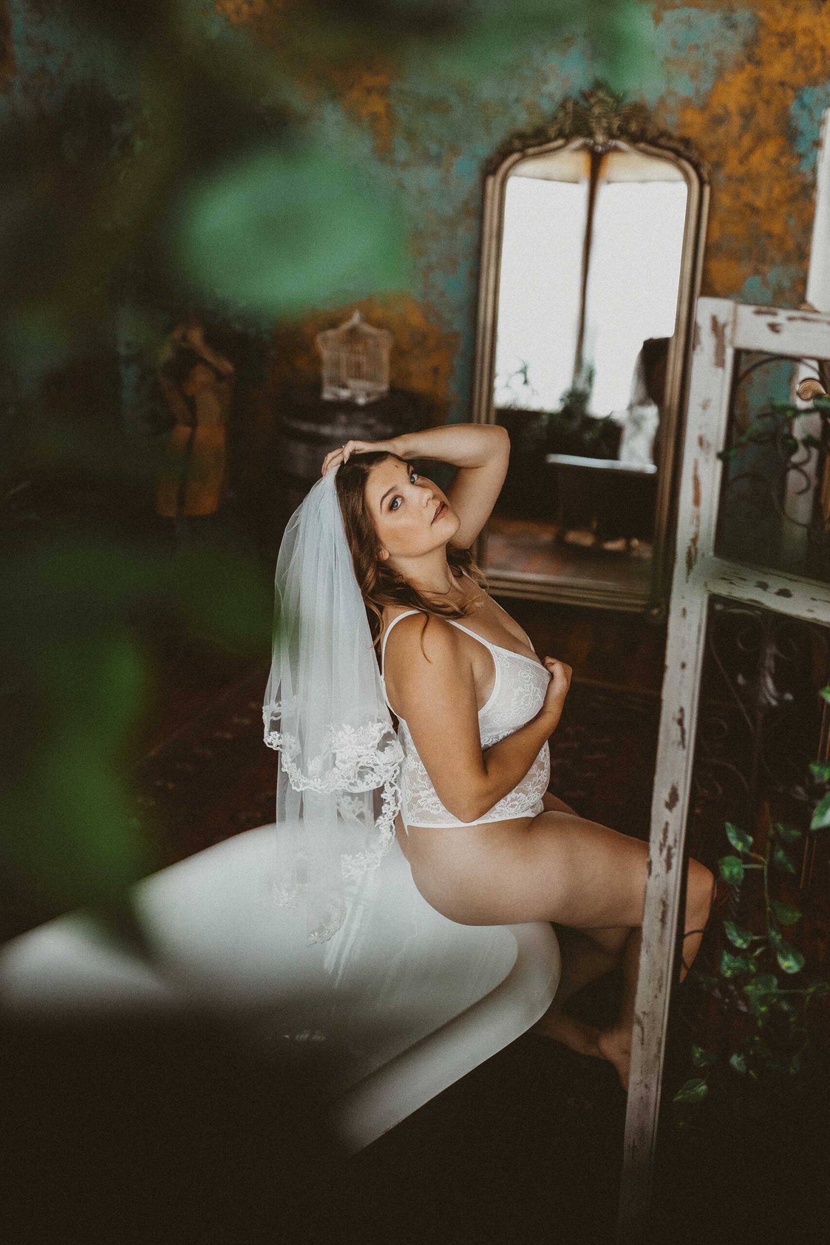 Bridal boudoir portrait of a woman with long brown hair wearing a veil and a white lace bodysuit while sitting on the side of a tub with plants around her in the Boudoir by J&S Photography studio near Boston, Massachusetts