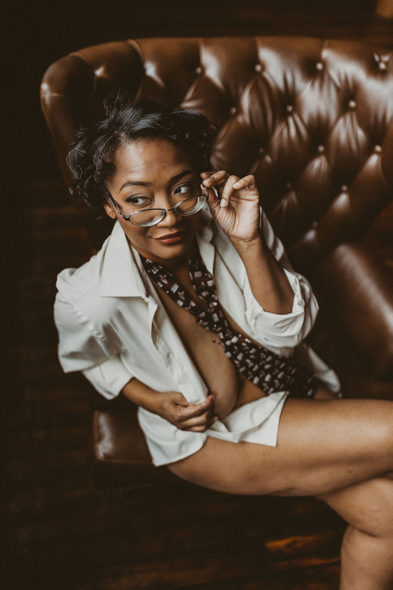 Boudoir portrait of a woman of color with short curly hair wearing glasses and a man's tie and button up white shirt while sitting cross legged in a brown leather armchair in the Boudoir by J&S Photography studio near Boston, Massachusetts