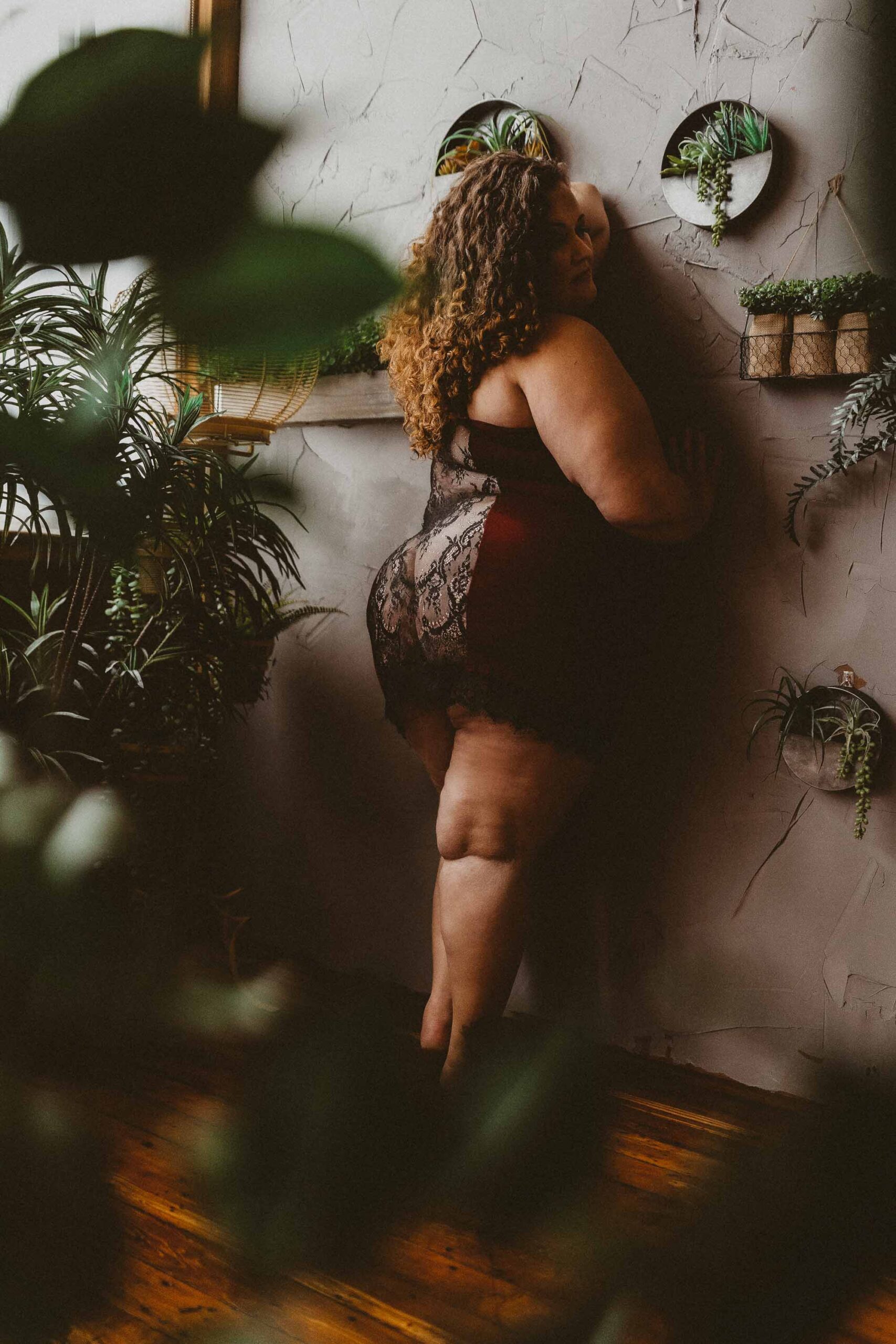 Curvy woman with long curly hair wearing a red and black lace dress facing a gray textured wall while surrounded by plants in a Boudoir photography studio