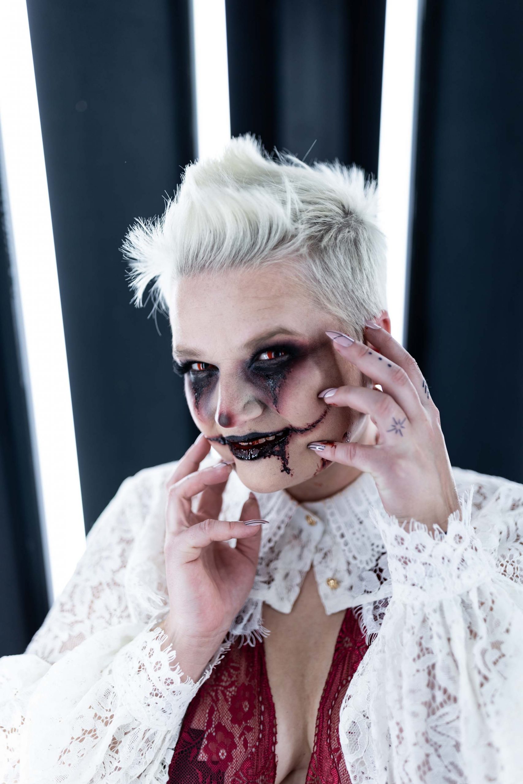 Jessie Clements wearing a red sheer lace bodysuit, white lace sleeves, and SFX demented harlequin cosplay makeup with red contacts in the Boudoir by J&S Photography studio near Boston, Massachusetts