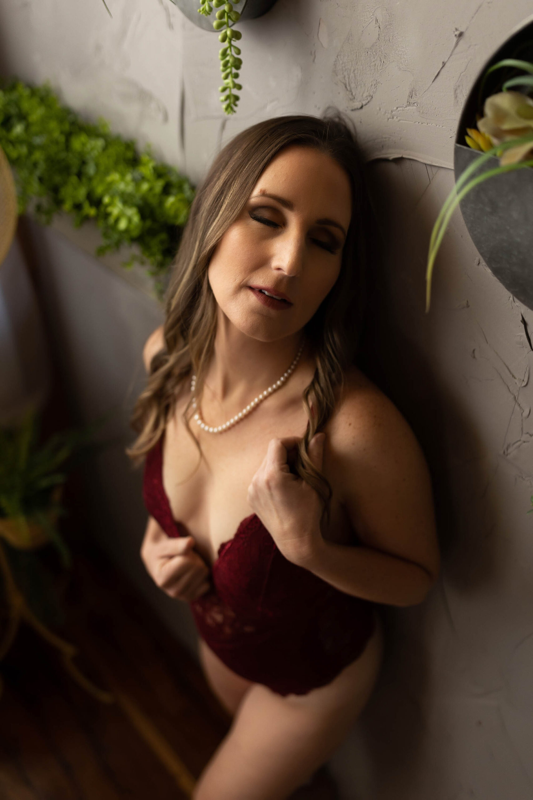 Brunette woman wearing a dark red lace bodysuit and pearls leaning against a gray textured wall with her eyes closed in the Boudoir by J&S Photography studio near Boston, Massachusetts