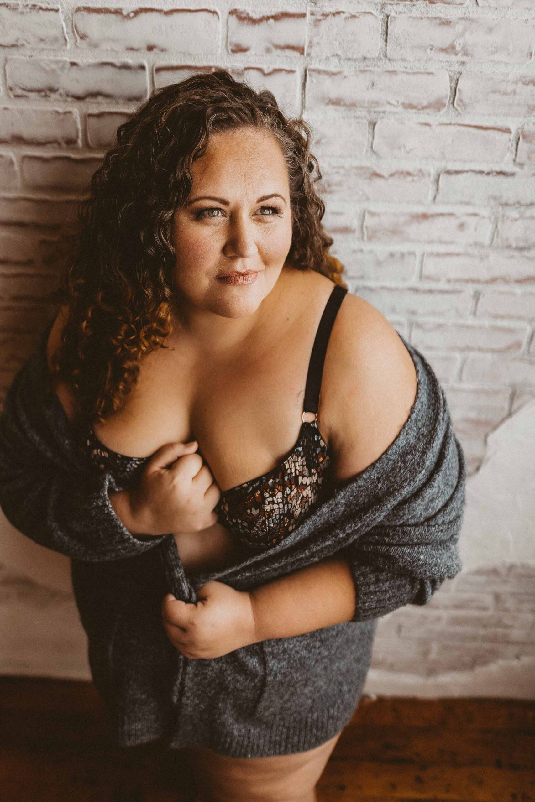 A plus size brunette woman with curly hair wearing a bra with a gray sweater draped around her giving a sultry look