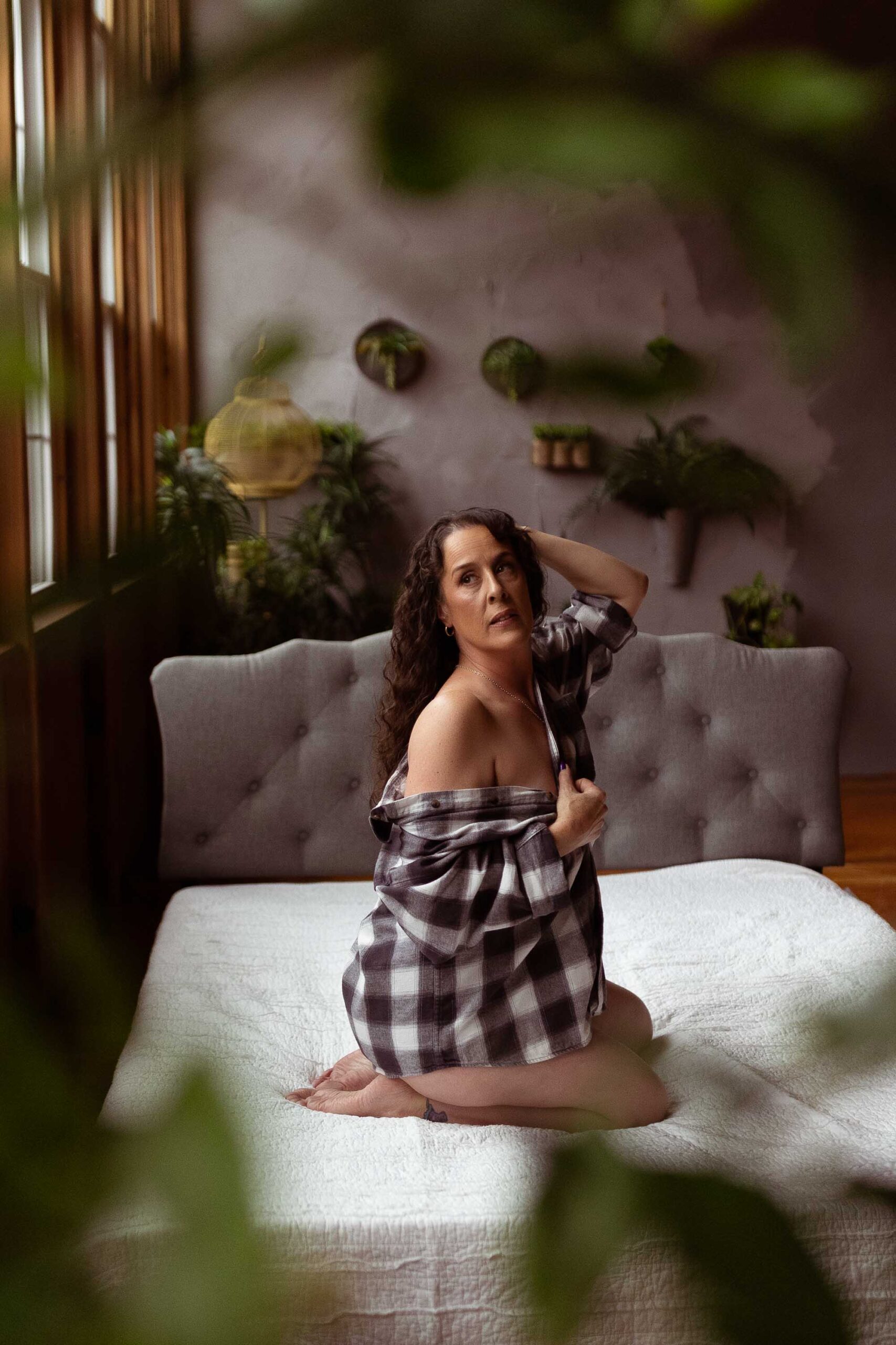 A brunette woman with curly hair kneels on a white bed with a flannel wrapped around her, looking out the windows behind her in the Boudoir by J&S Photography studio near Boston, Massachusetts
