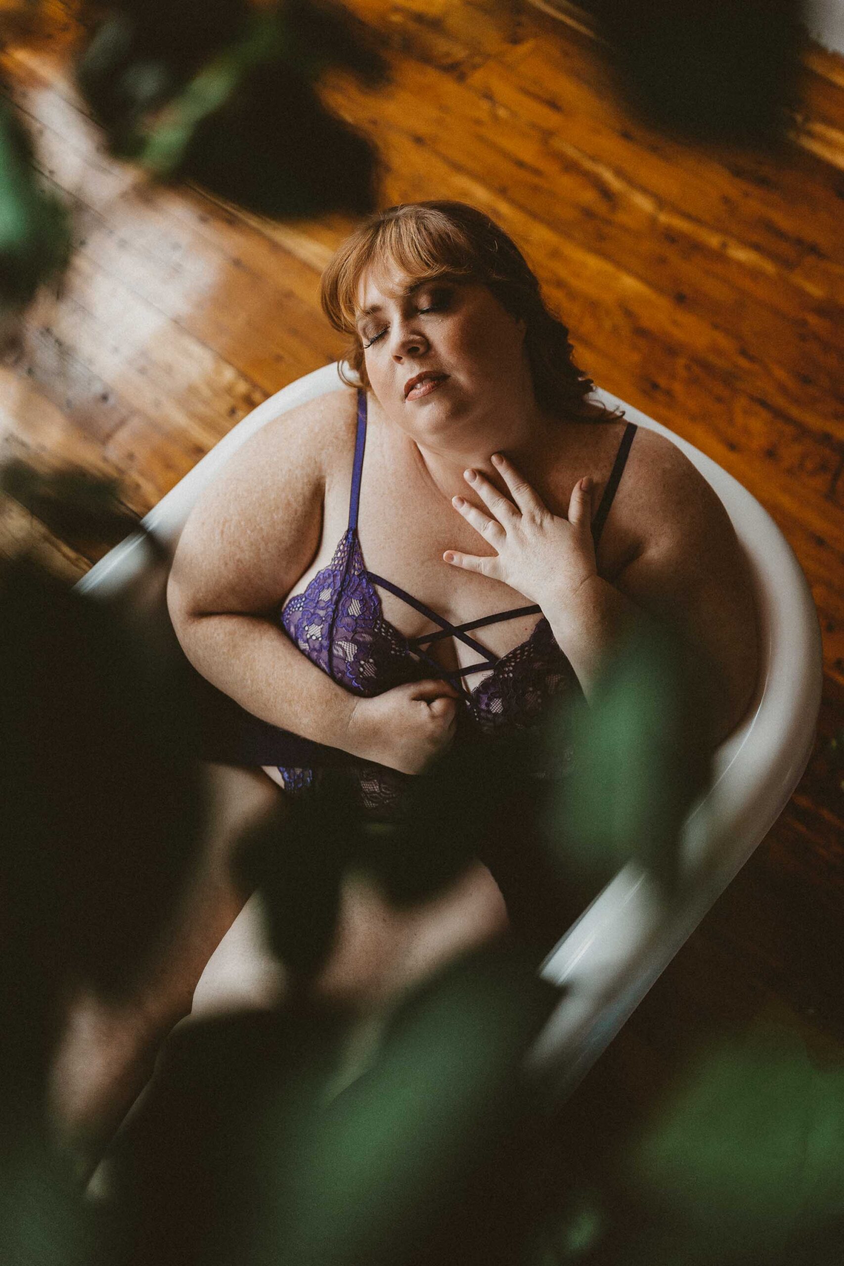 A plus size woman with red hair feeling blissful, laying back in a bathtub wearing purple lace lingerie in the Boudoir by J&S Photography studio near Boston, Massachusetts