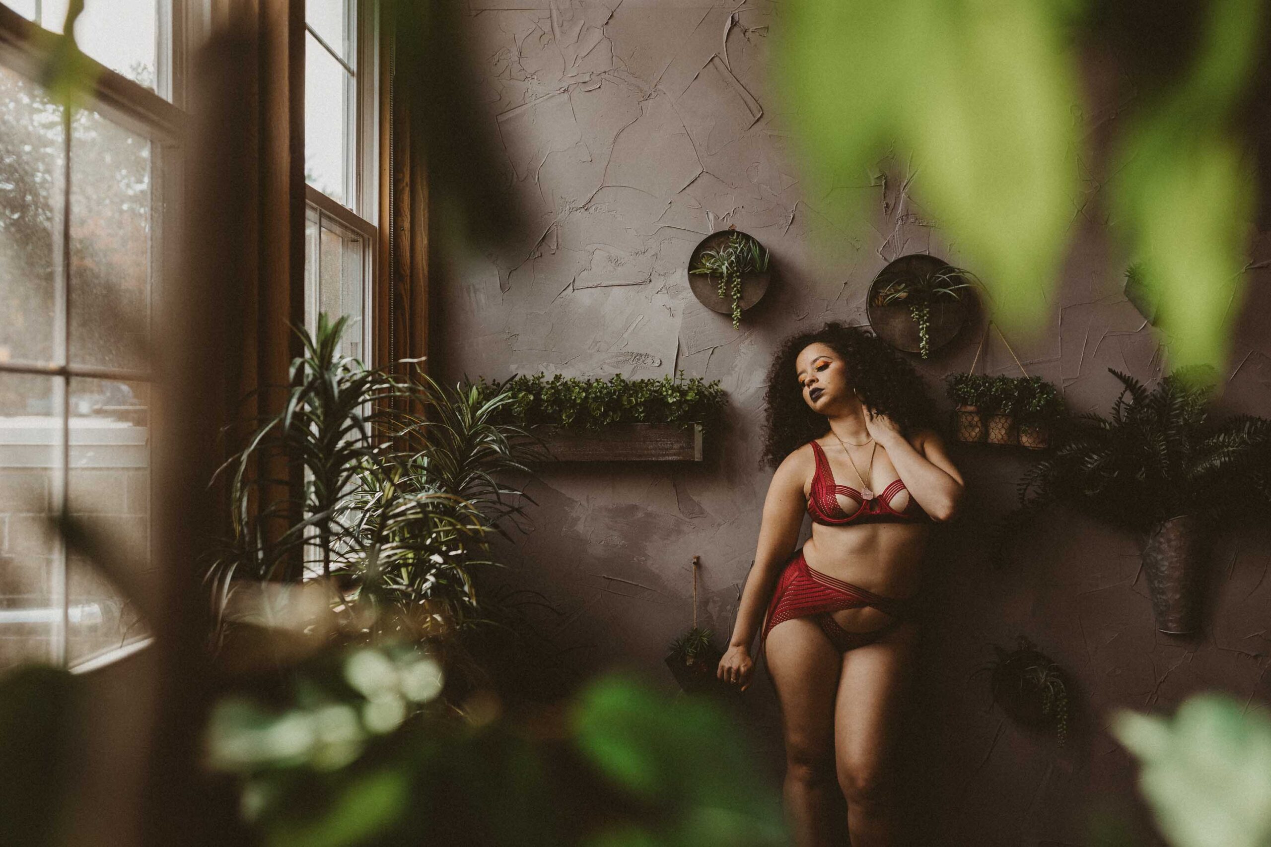 A woman of color wearing a red lingerie set stands in a photography studio surrounded by plants in Massachusetts.
