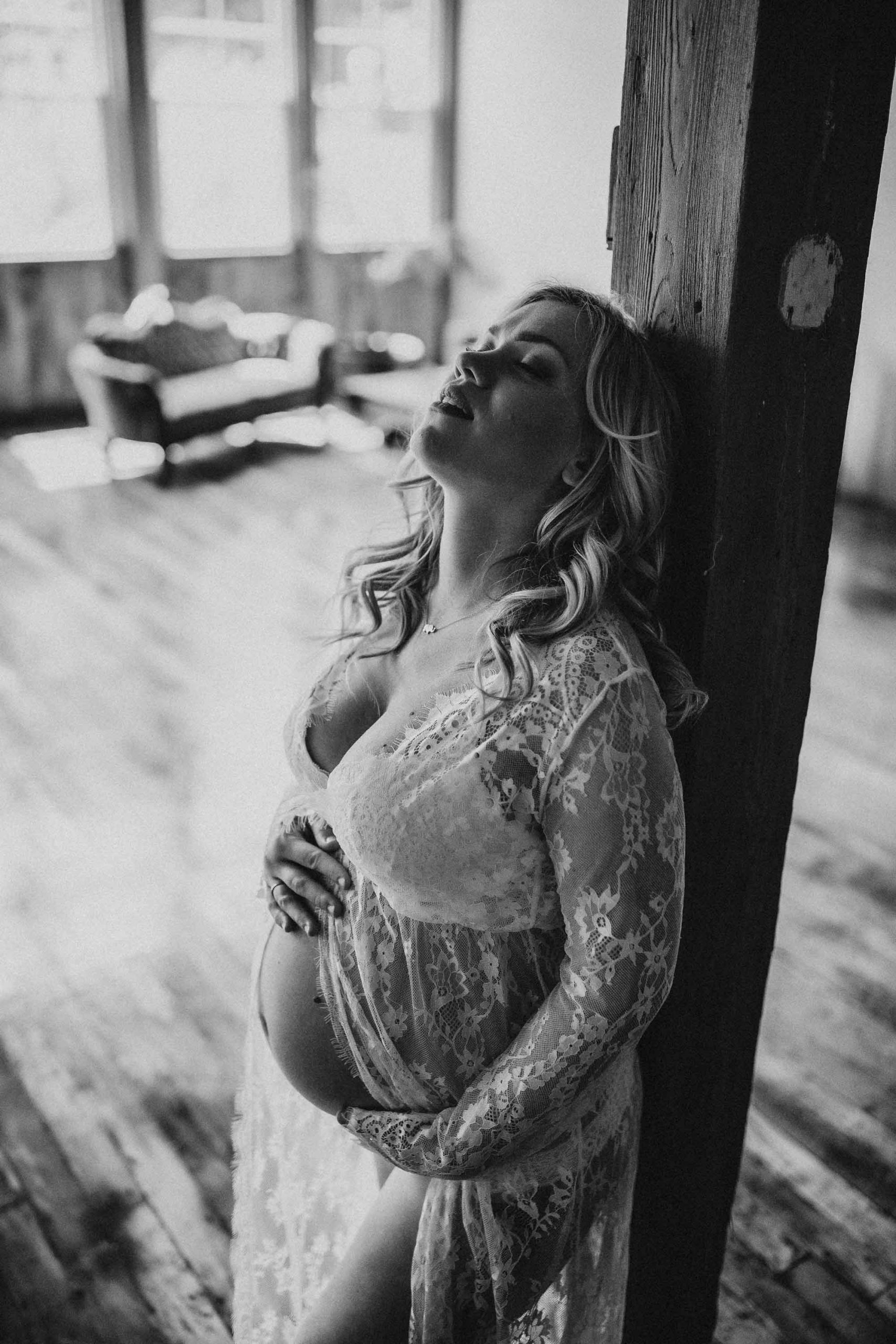 A pregnant woman waring a white lace dress leans against a pillar with her eyes closed in the Boudoir by J&S Photography studio near Boston, Massachusetts.