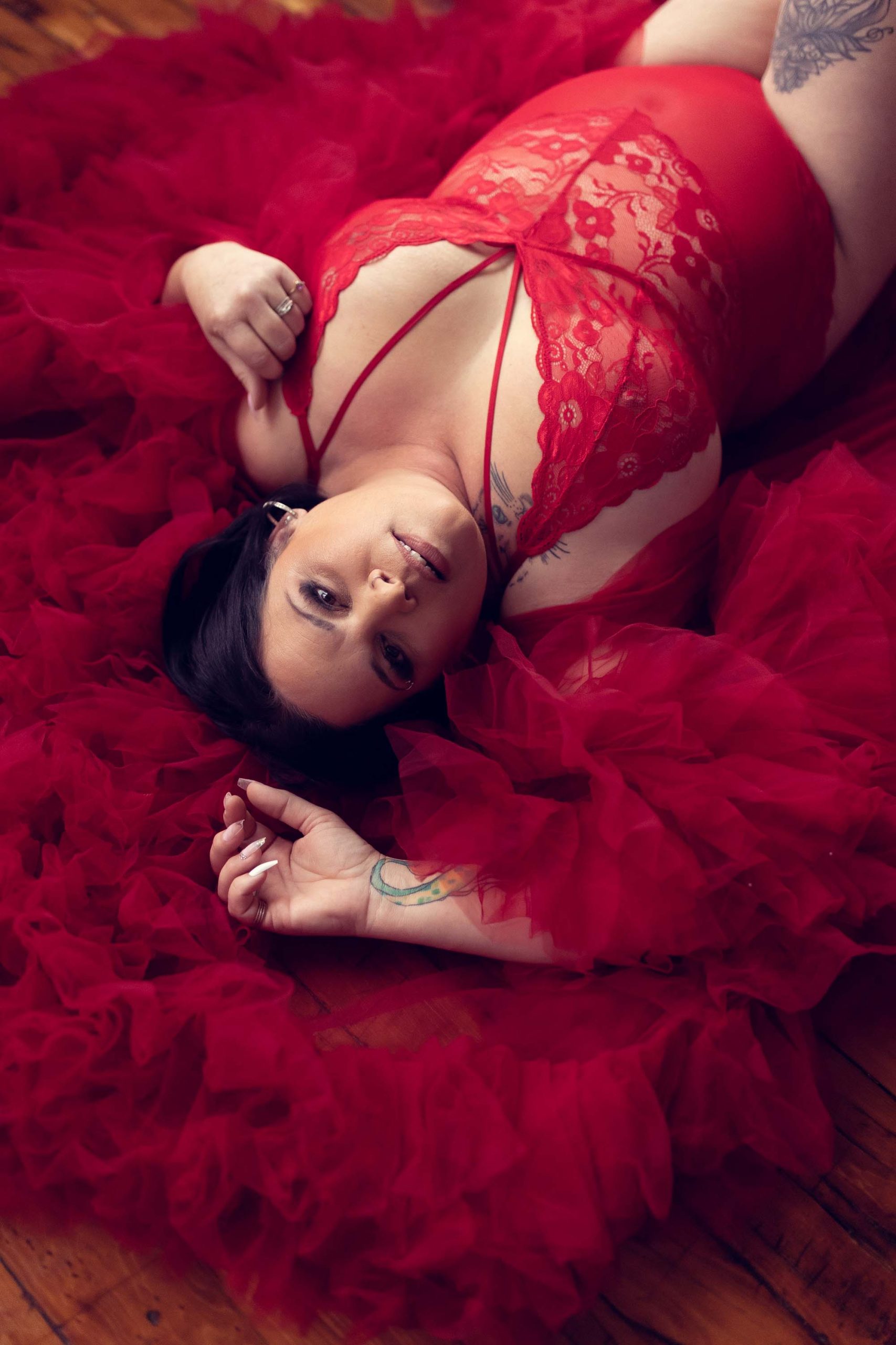 A woman with tattoos wearing a red bodysuit and ruffled robe reclines on a hardwood floor in a boudoir photography studio.